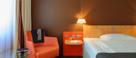A hotel room with a bed, a glass table, an orange chair, and a nightstand with a lamp.