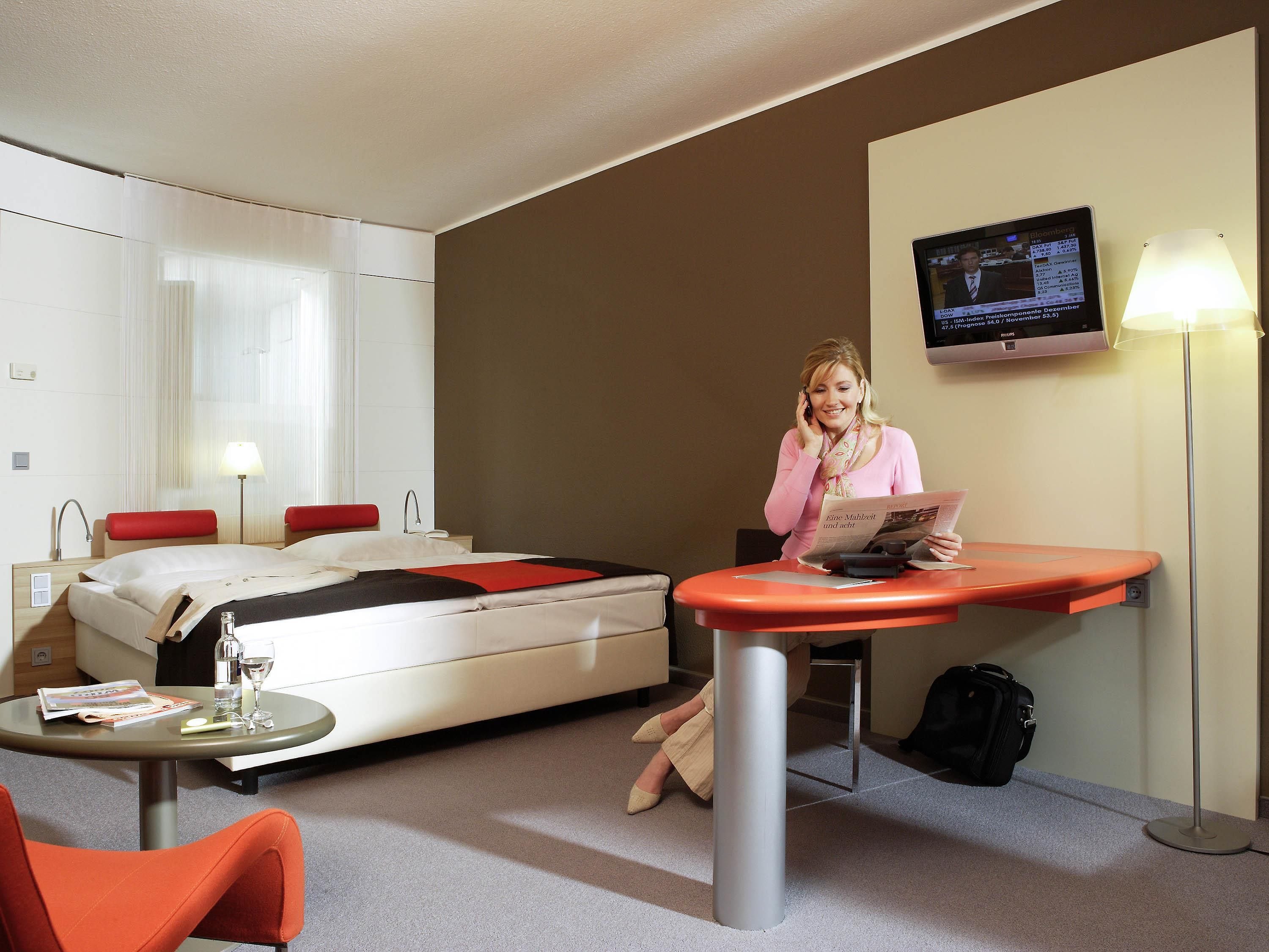 A hotel room with a bed, a desk, a chair, a TV, and a lamp.