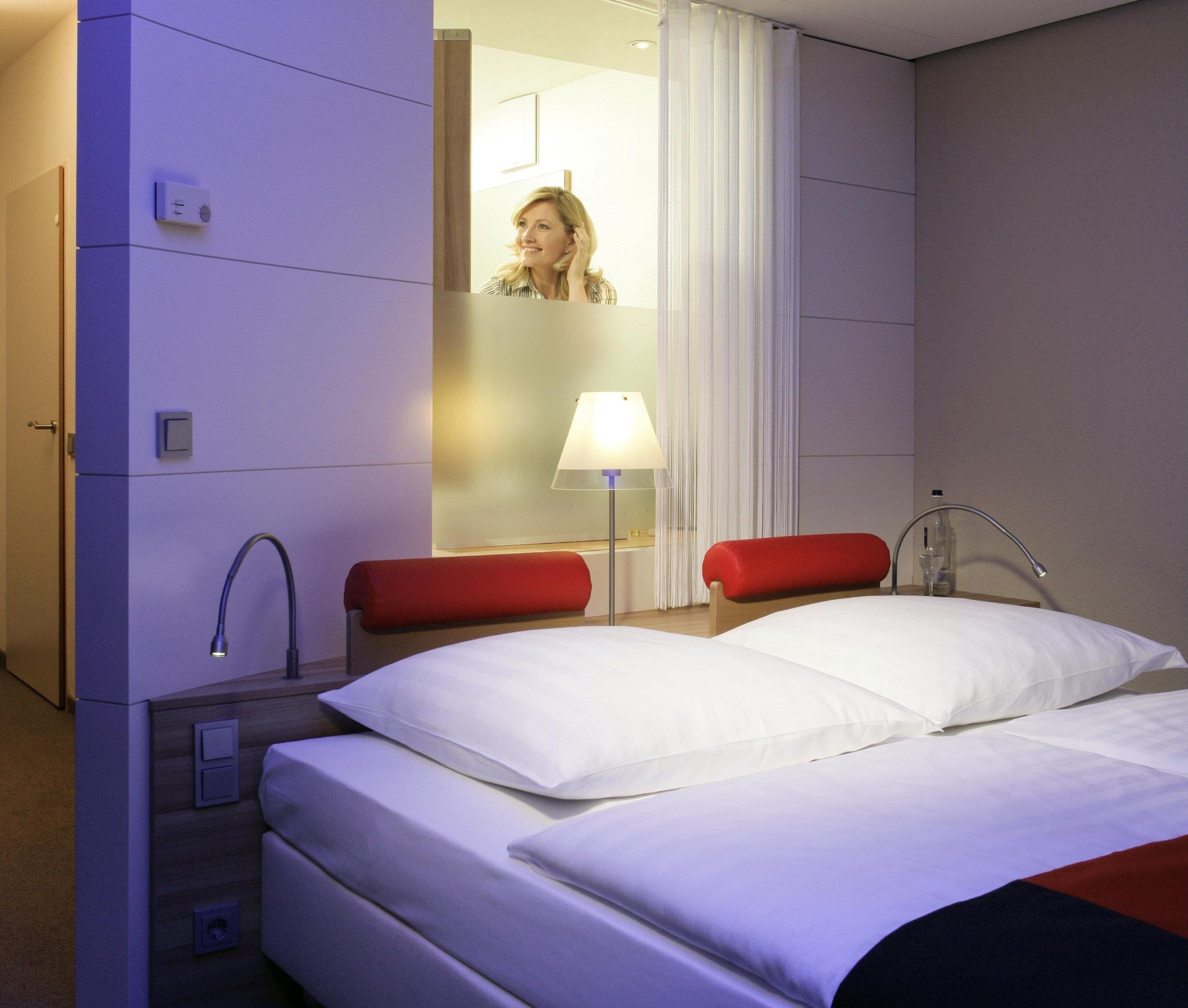 A hotel room with a bed, two red chairs, a nightstand, a lamp, and a mirror.
