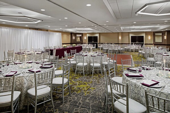 Un salón de banquetes con mesas redondas, sillas blancas y una zona escenario.