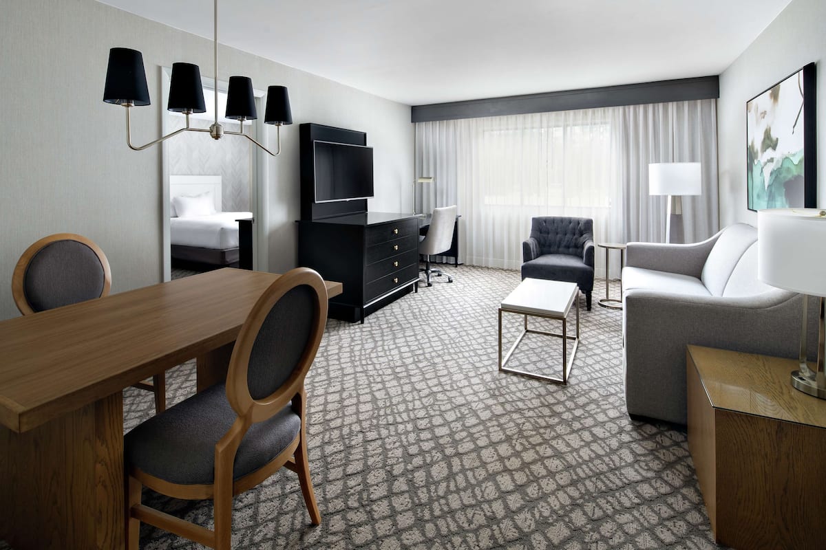Una habitación de hotel con una mesa de comedor de madera, dos sillas, un mueble negro para televisión, un sofá gris, un sillón blanco y una alfombra estampada.