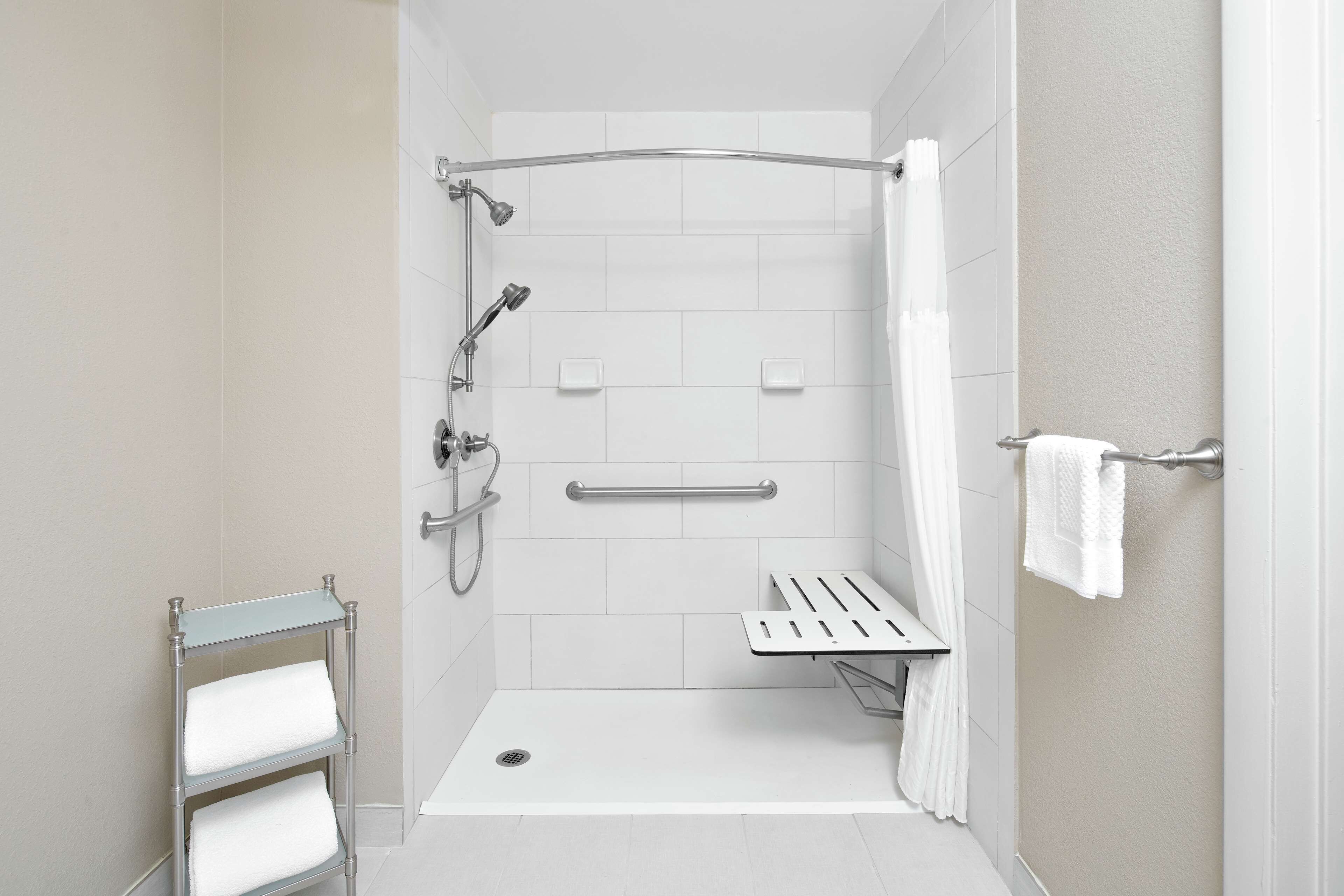 A modern shower area with a glass door, a handrail, a bench, and a towel rack.