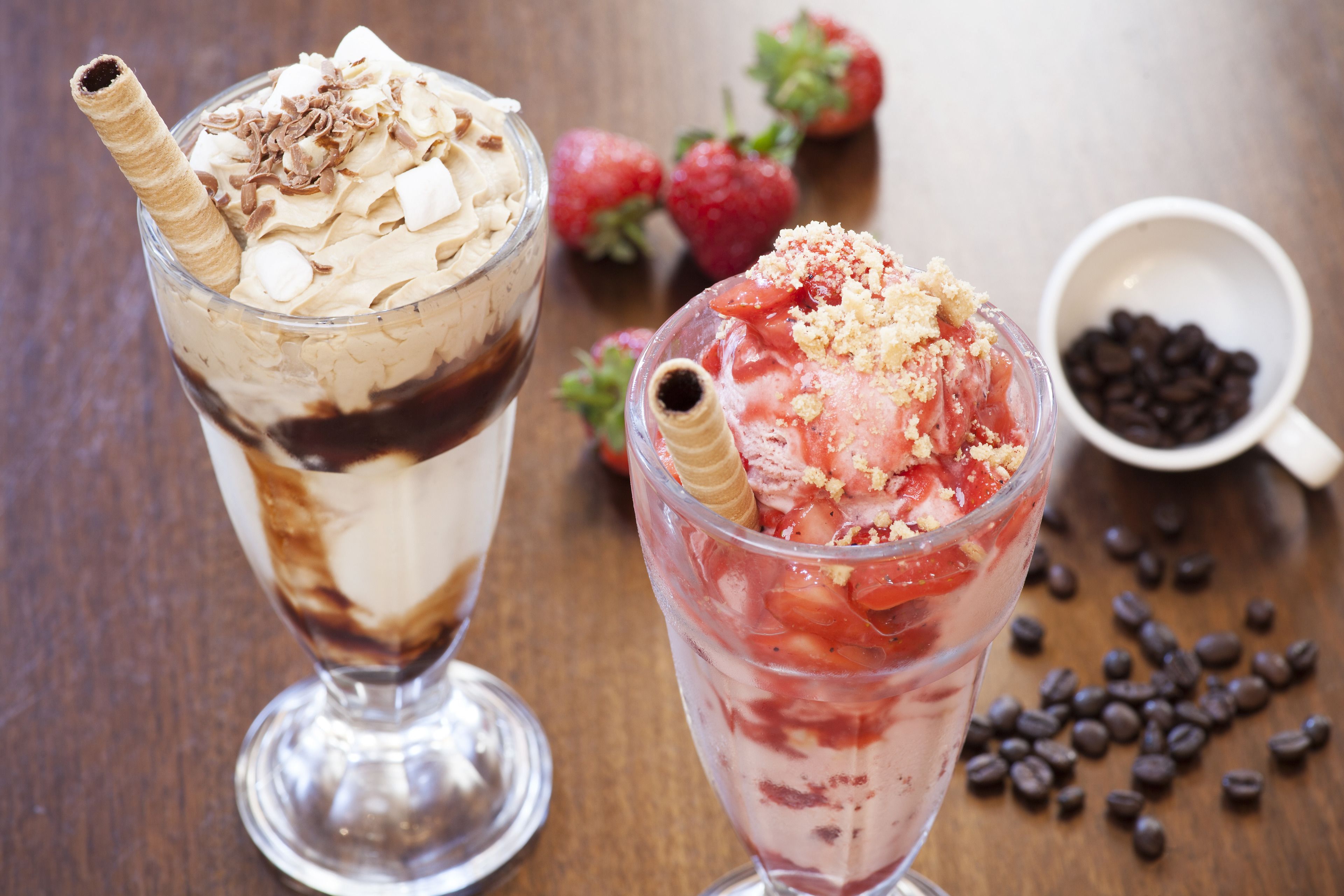 Two layered desserts in clear glasses, strawberries, and a bowl of chocolate chips on a wooden table.