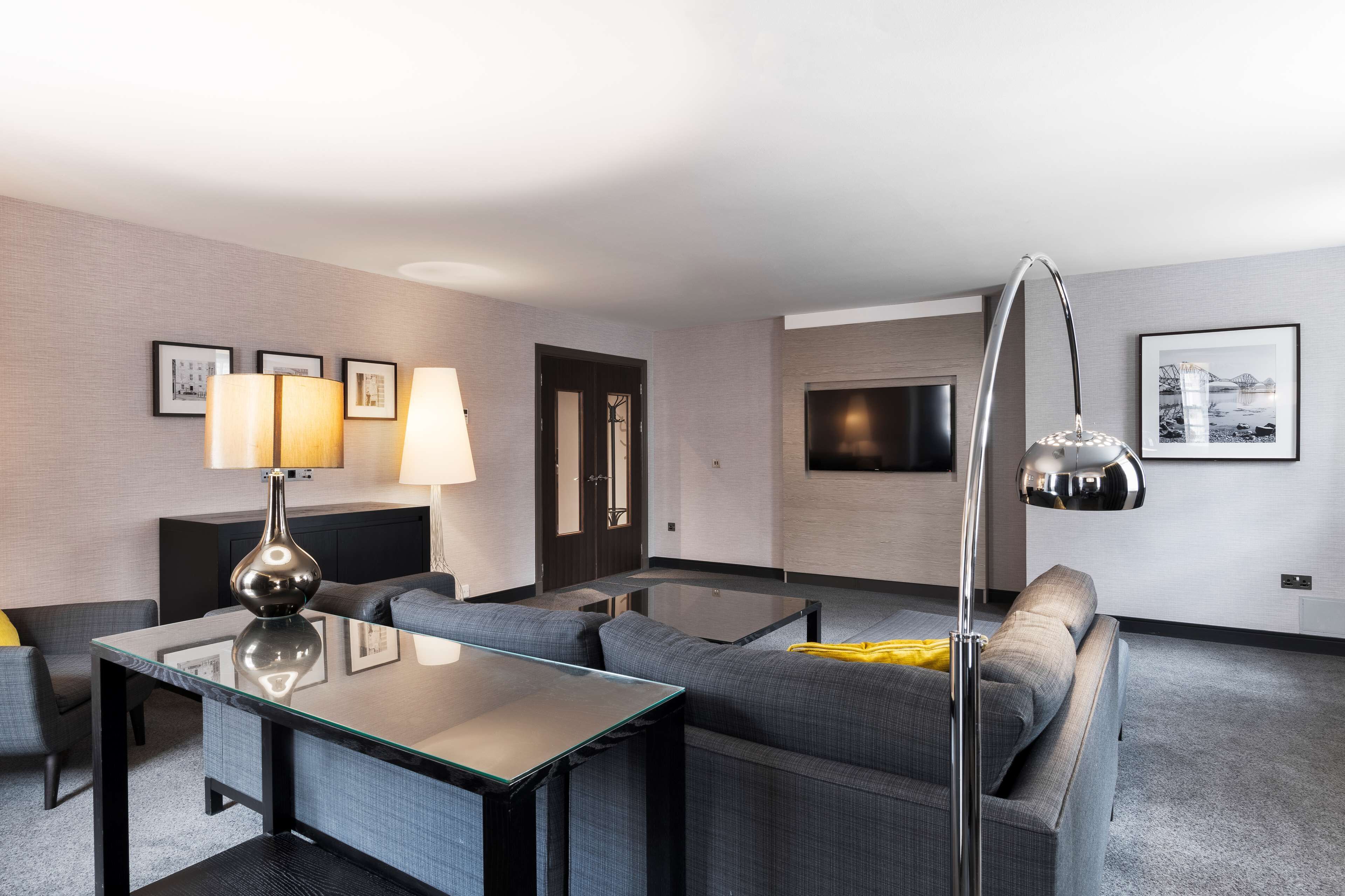 A modern hotel room with a glass coffee table, grey sofa, and a TV mounted on the wall.