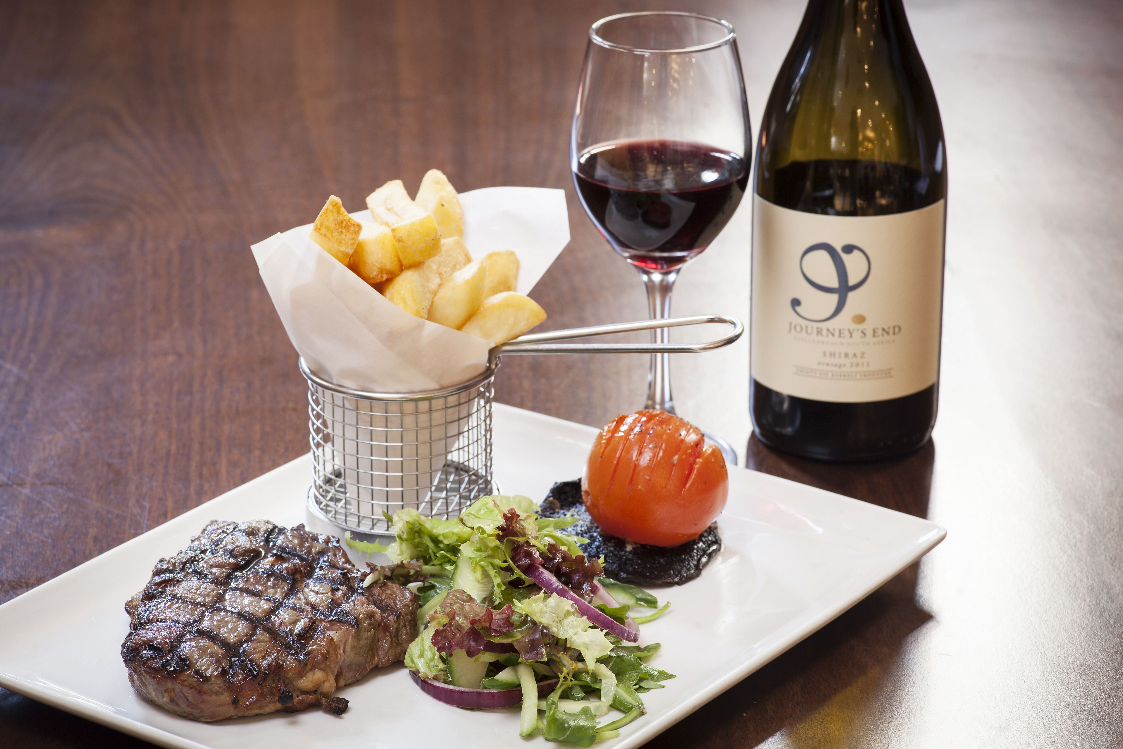 Un plato con bistec a la parrilla, ensalada, papas asadas y una copa de vino tinto.