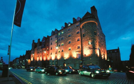 A historic stone building with multiple windows and a turret, illuminated by warm lights against a twilight sky.