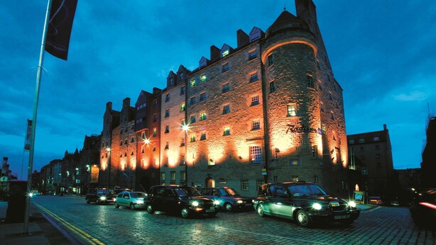 A historic stone building with multiple windows and a turret, illuminated by warm lights against a twilight sky. - Radisson Blu Hotel, Edinburgh City Centre (Edinburgh)