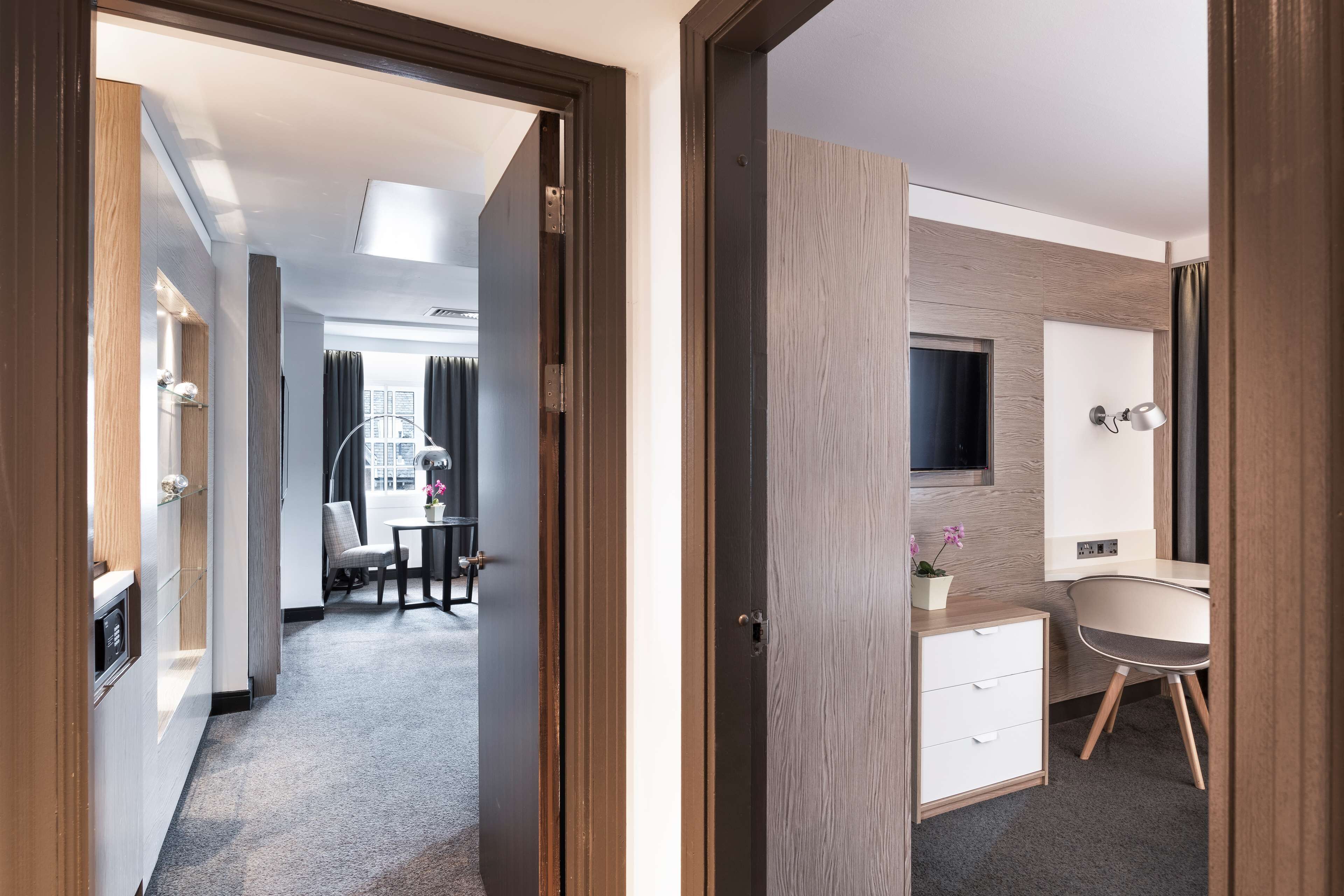 A hotel room with a wooden door, a desk with a chair, a small bathroom with a mirror, and a TV mounted on the wall.