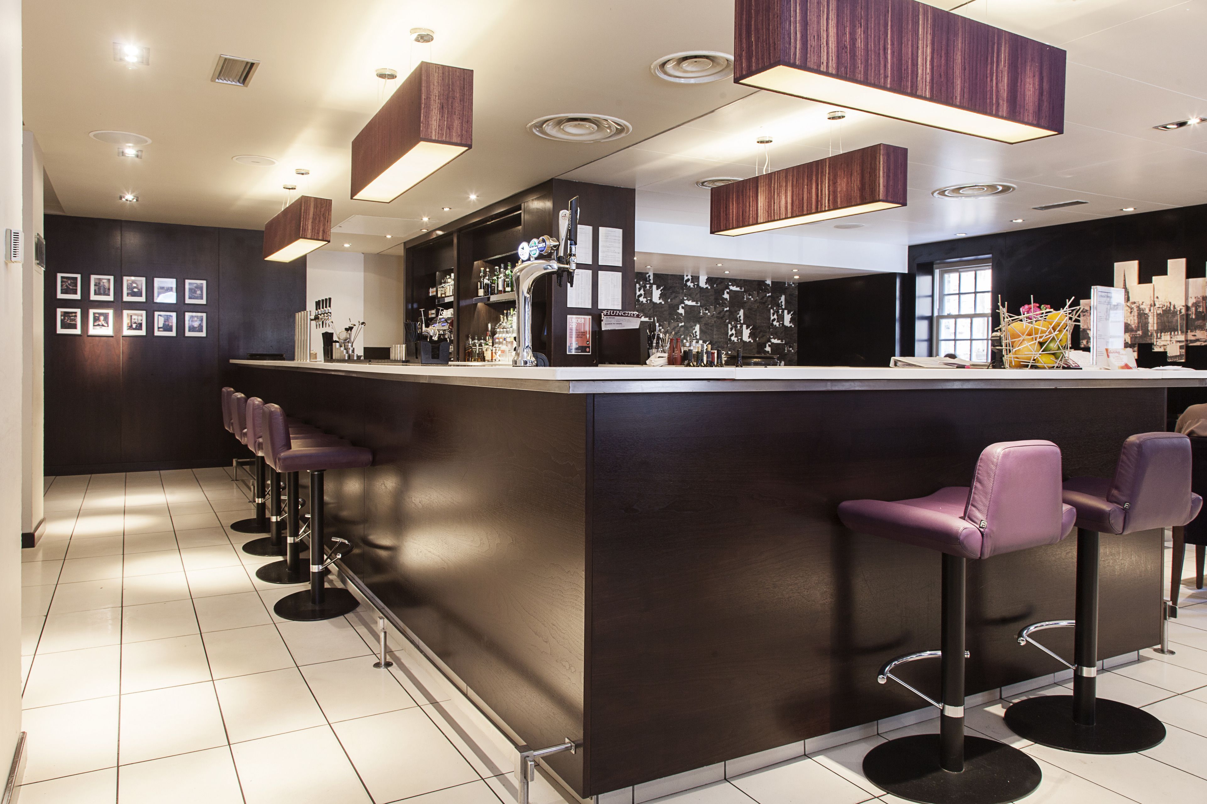A modern bar area with a dark wood bar counter, purple bar stools, and overhead lighting.