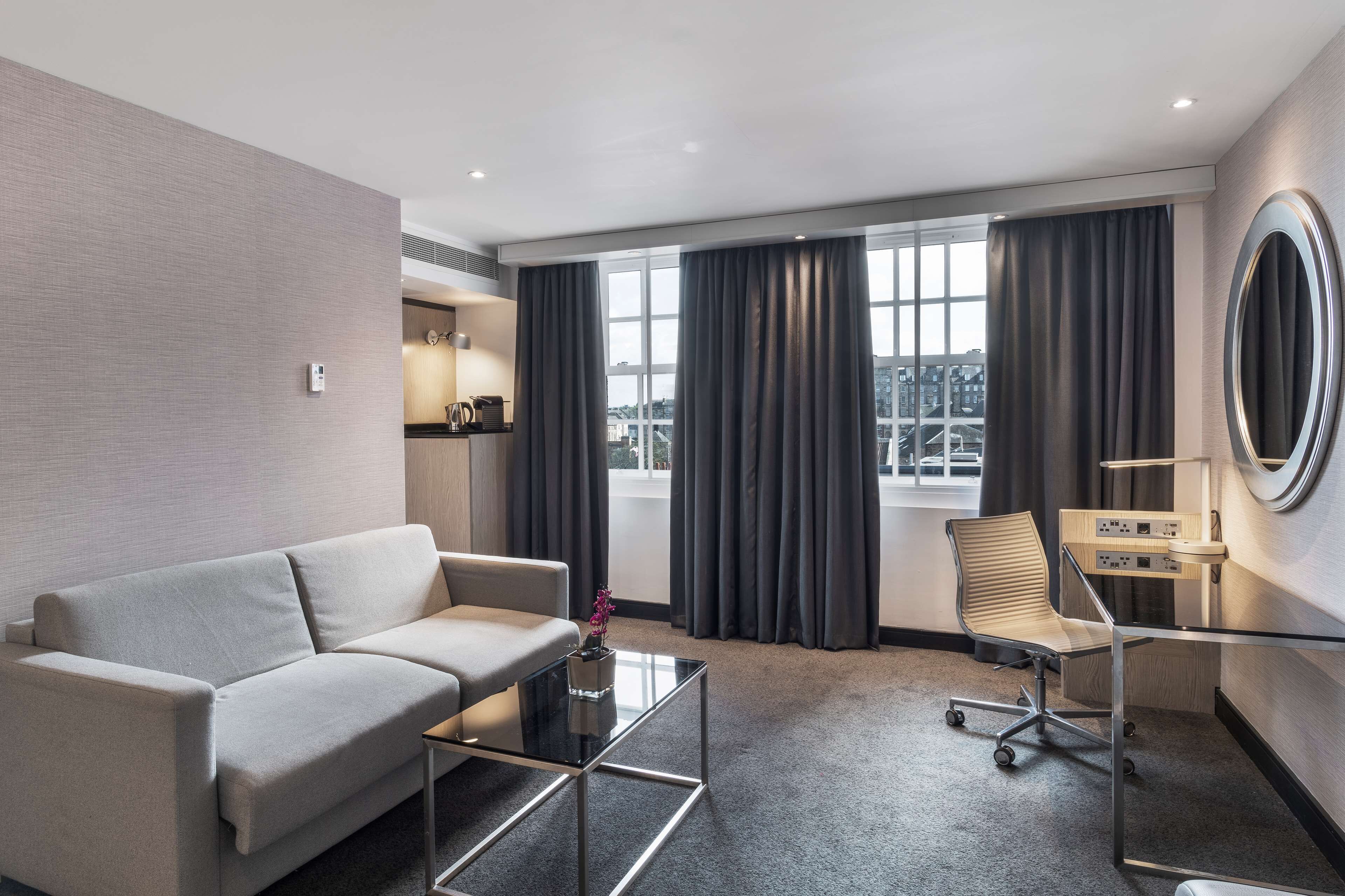 A modern hotel room with a sofa, a glass coffee table, a desk with a chair, a large window with curtains, and a view of the city.
