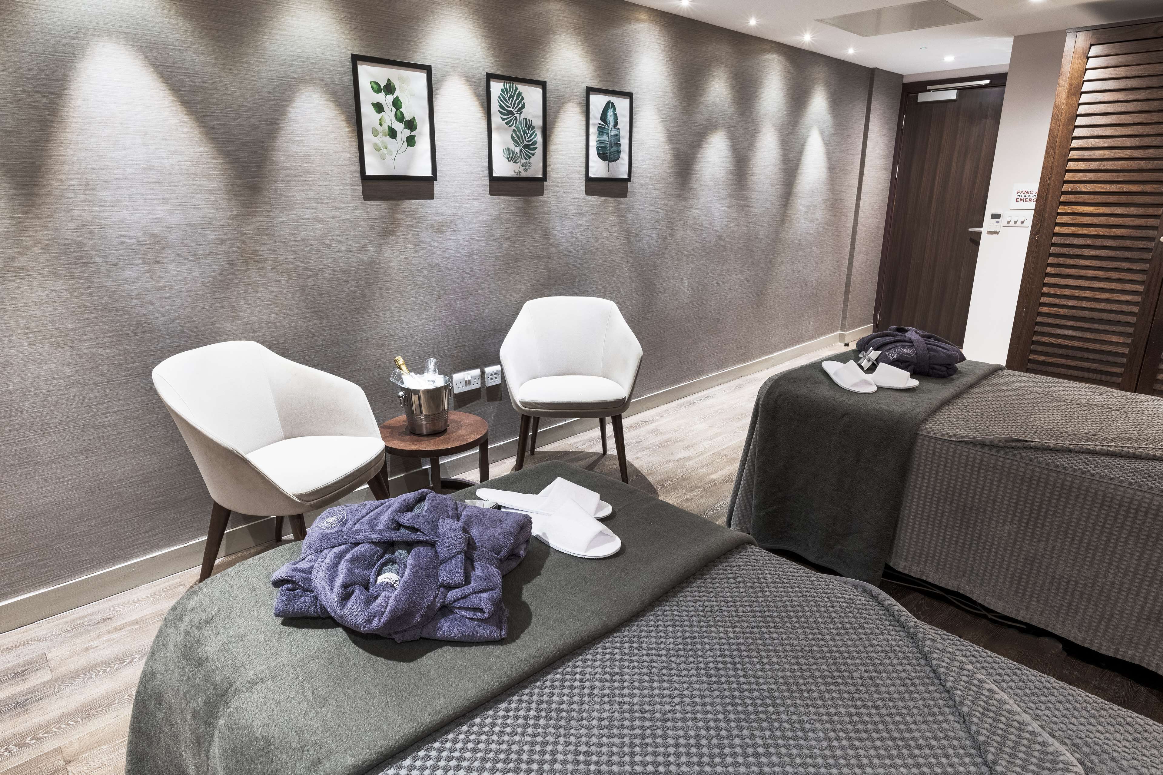A hotel room with two white armchairs, a small table with a lamp, a bed with a grey blanket, and a wall with framed leaf art.