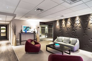 A modern lobby with a sofa, armchair, and coffee table, a reception desk, and a decorative wall with leaf patterns.