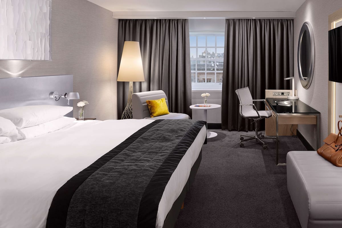 Une chambre d’hôtel moderne, dotée d’un grand lit, d’un bureau, d’une chaise, d’une petite table avec une lampe, et offrant une vue sur la ville par la fenêtre.