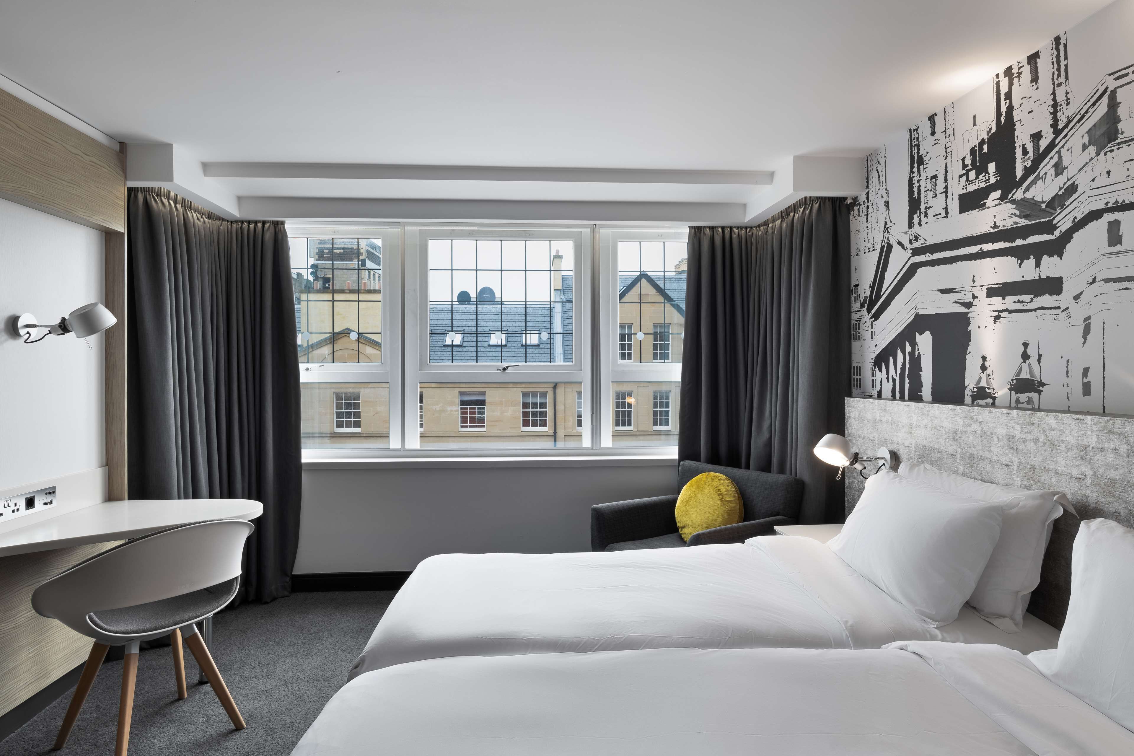 Habitación de hotel con una cama, un escritorio y una silla. Hay una ventana con cortinas, una lámpara y una pared con un mural de un paisaje urbano.