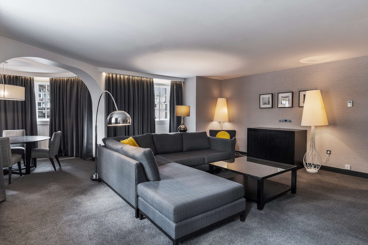 Une chambre d’hôtel moderne avec un canapé d’angle gris, une petite table ronde et deux lampadaires.