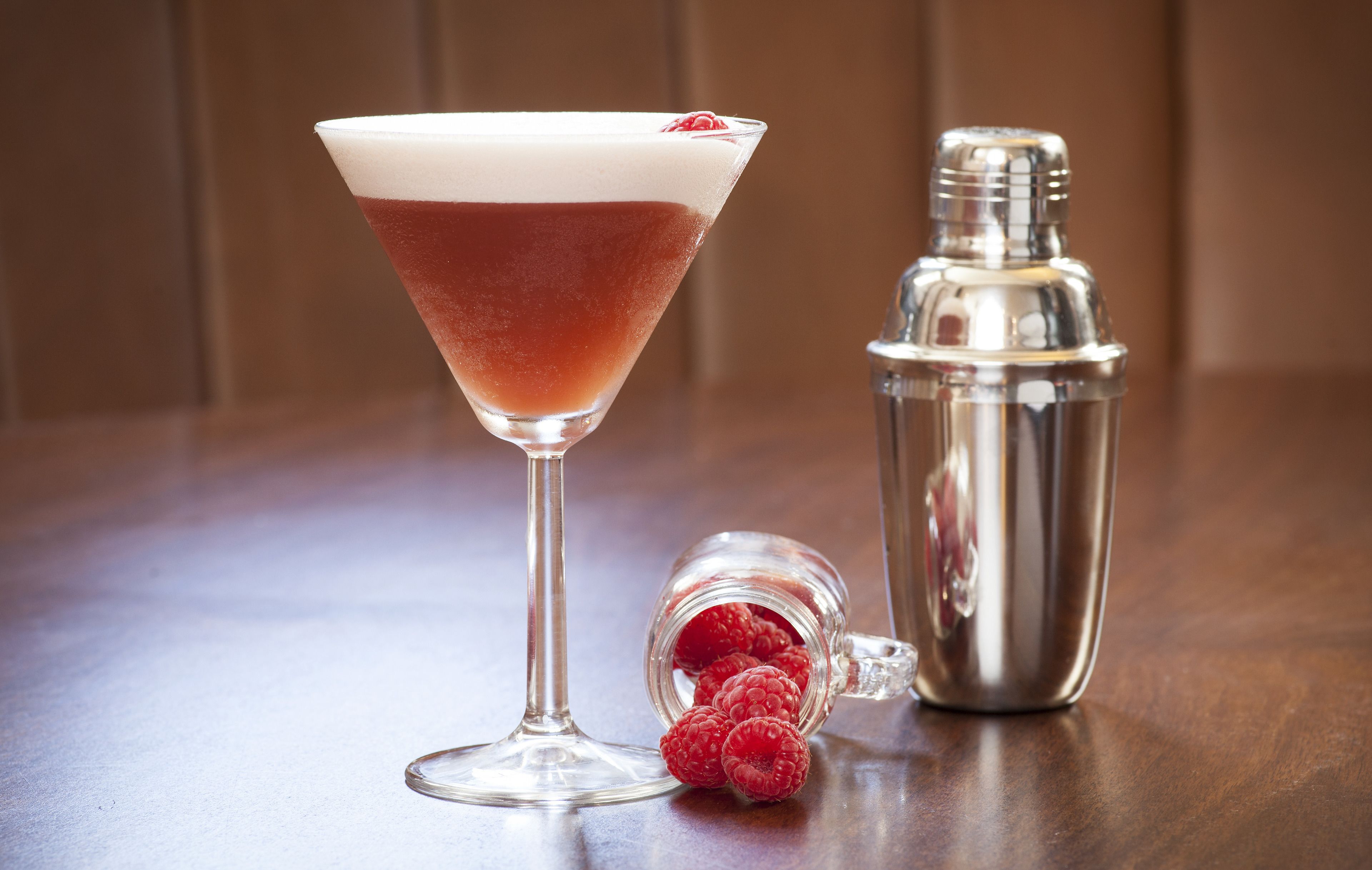 A martini glass with a layered drink, a cocktail shaker, and a jar of raspberries on a wooden surface.