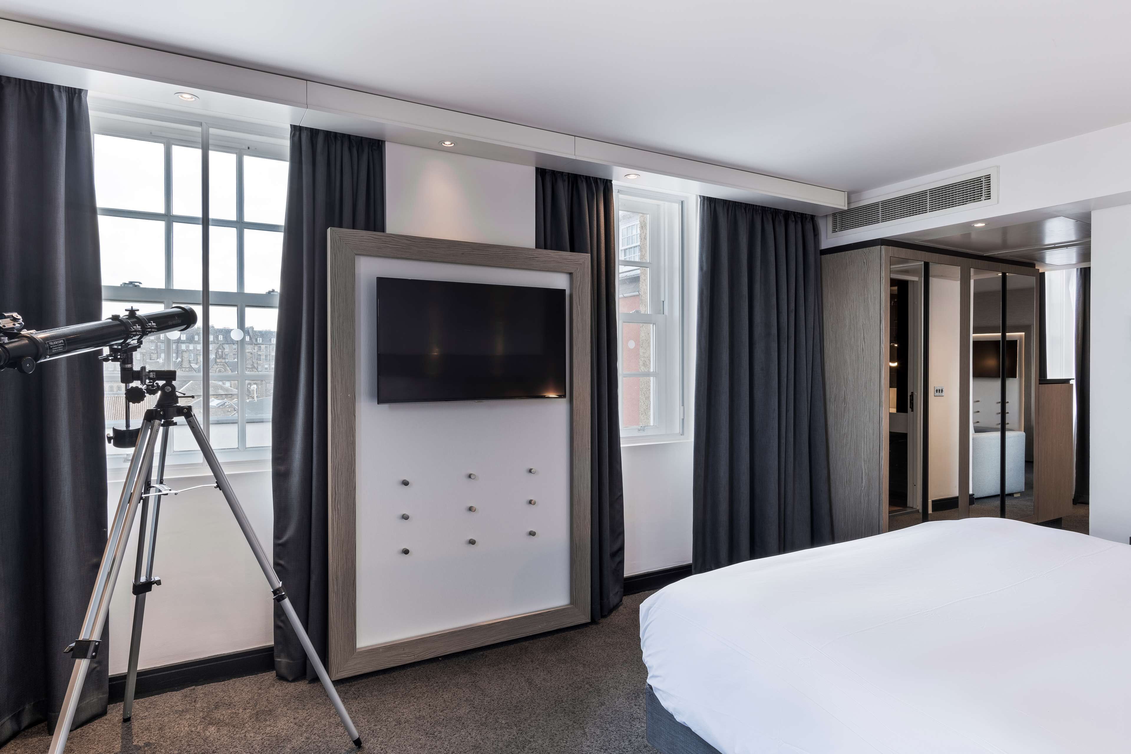 A hotel room with a large bed, a mounted telescope, a flat-screen TV, and a view of the city through large windows.