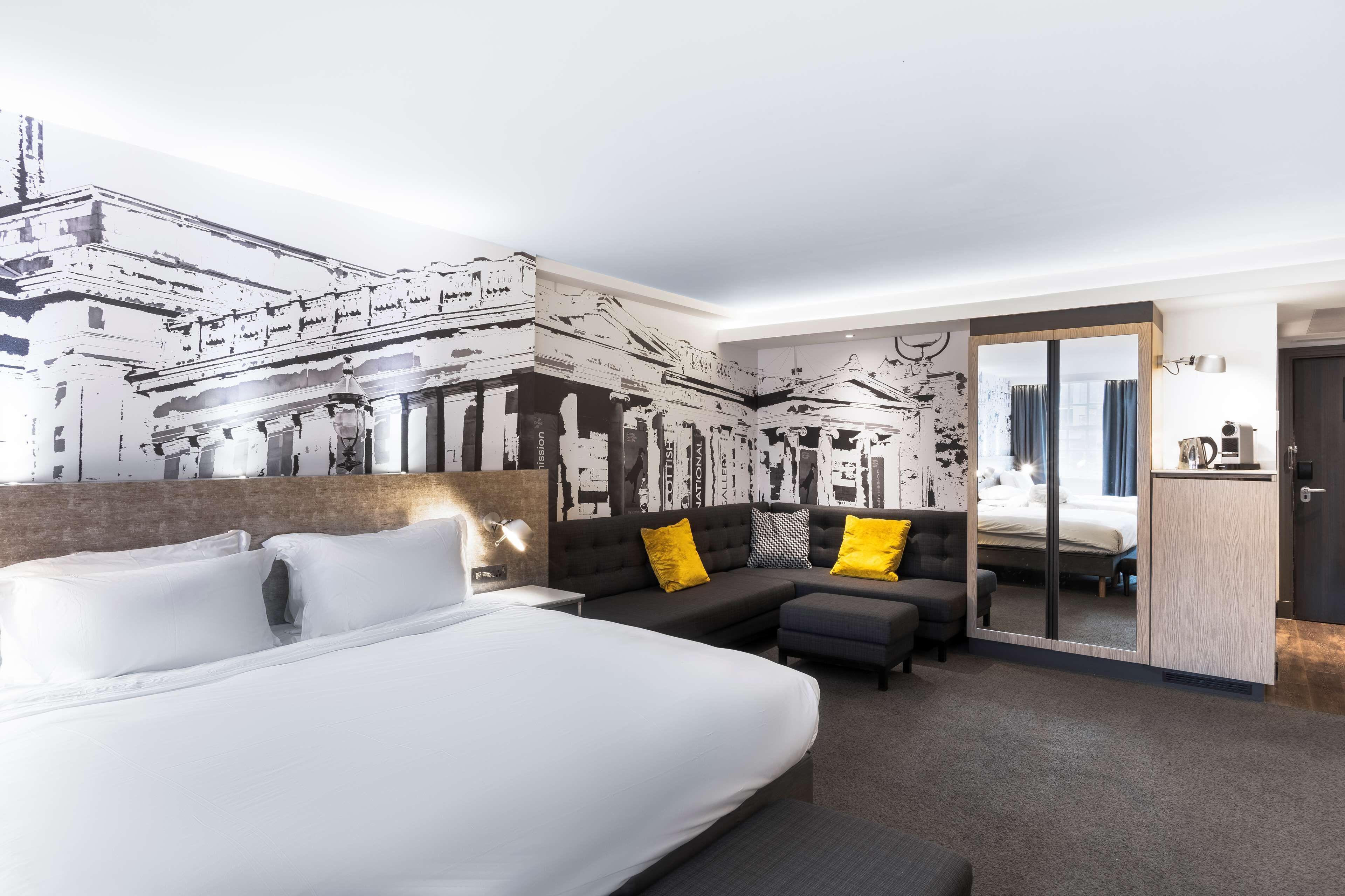 A modern hotel room with a large bed, a sofa, a small table, and a mirror.