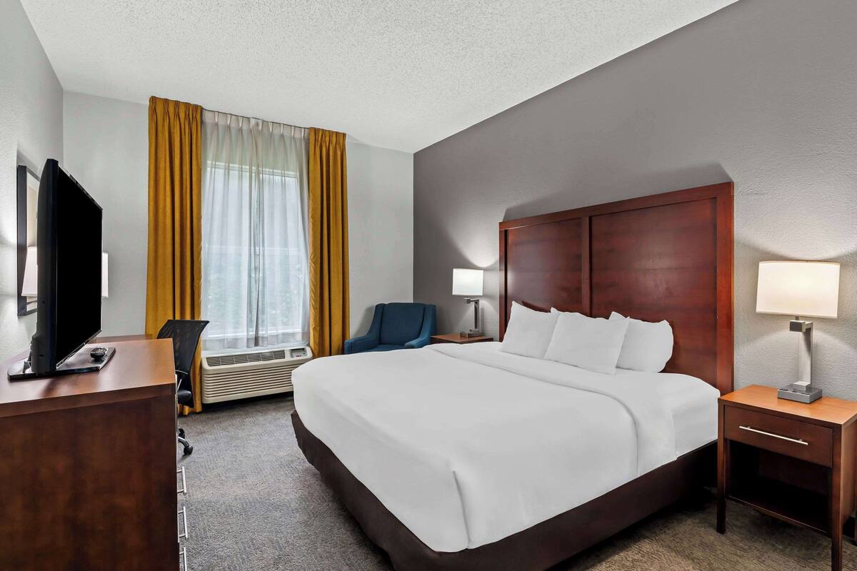 A hotel room with a large bed, a desk with a flat-screen TV, a chair, a bedside table with a lamp, and a window with curtains.
