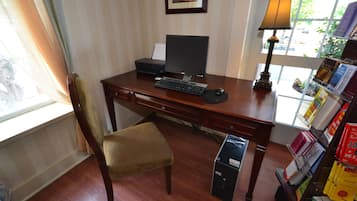A wooden desk with a computer, a lamp, and a chair. There is a bookshelf with books and a window with curtains.