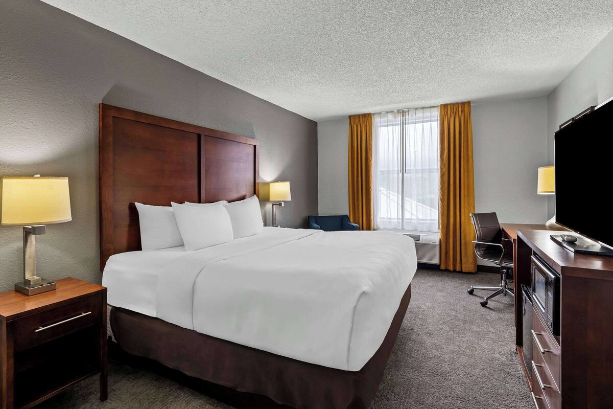 A hotel room with a large bed, a desk with a lamp, a chair, a television, and a window with curtains.