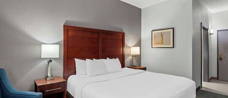 A hotel room with a large bed, a wooden headboard, a blue armchair, a nightstand with a lamp, and a framed picture on the wall.