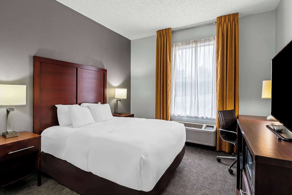 A hotel room with a large bed, a desk with a computer, a TV, and a window with curtains.