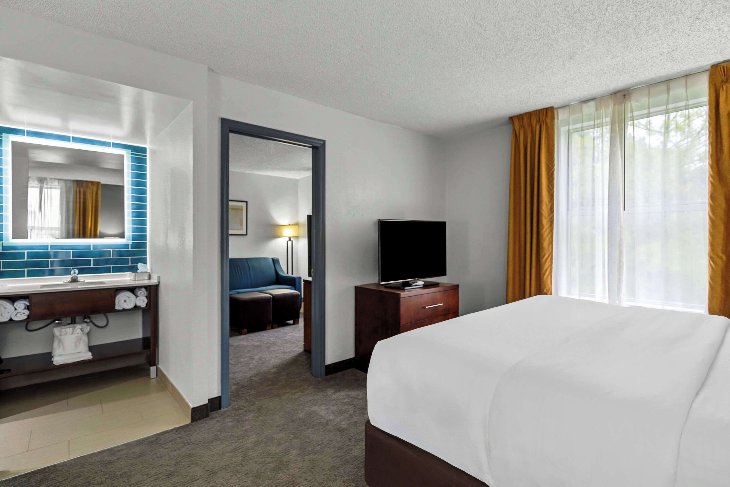 Habitación de hotel con cama, televisión, escritorio, silla, baño con espejo y ventana con cortinas.