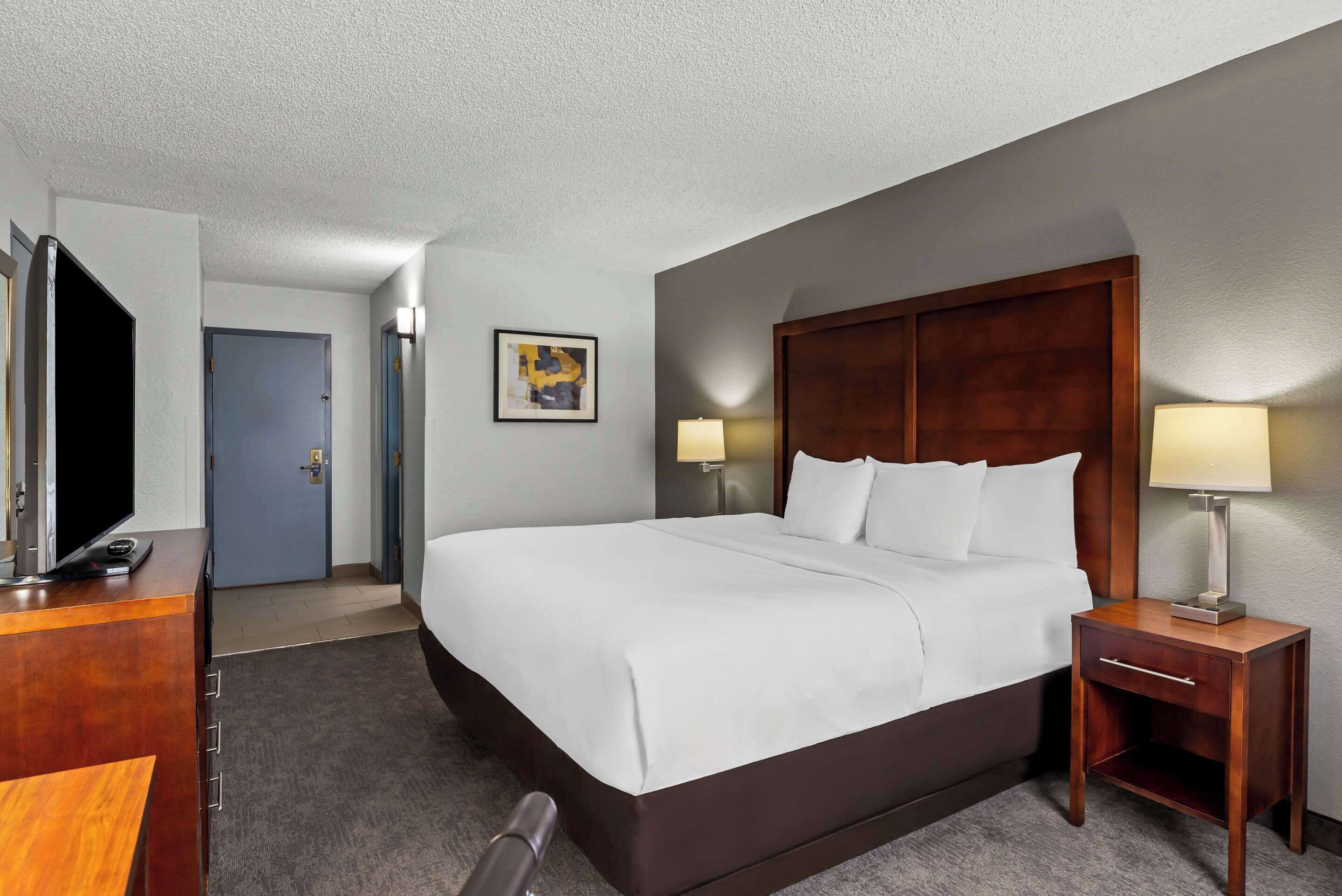 A hotel room with a large bed, a desk with a TV, a wooden nightstand with a lamp, and a door.