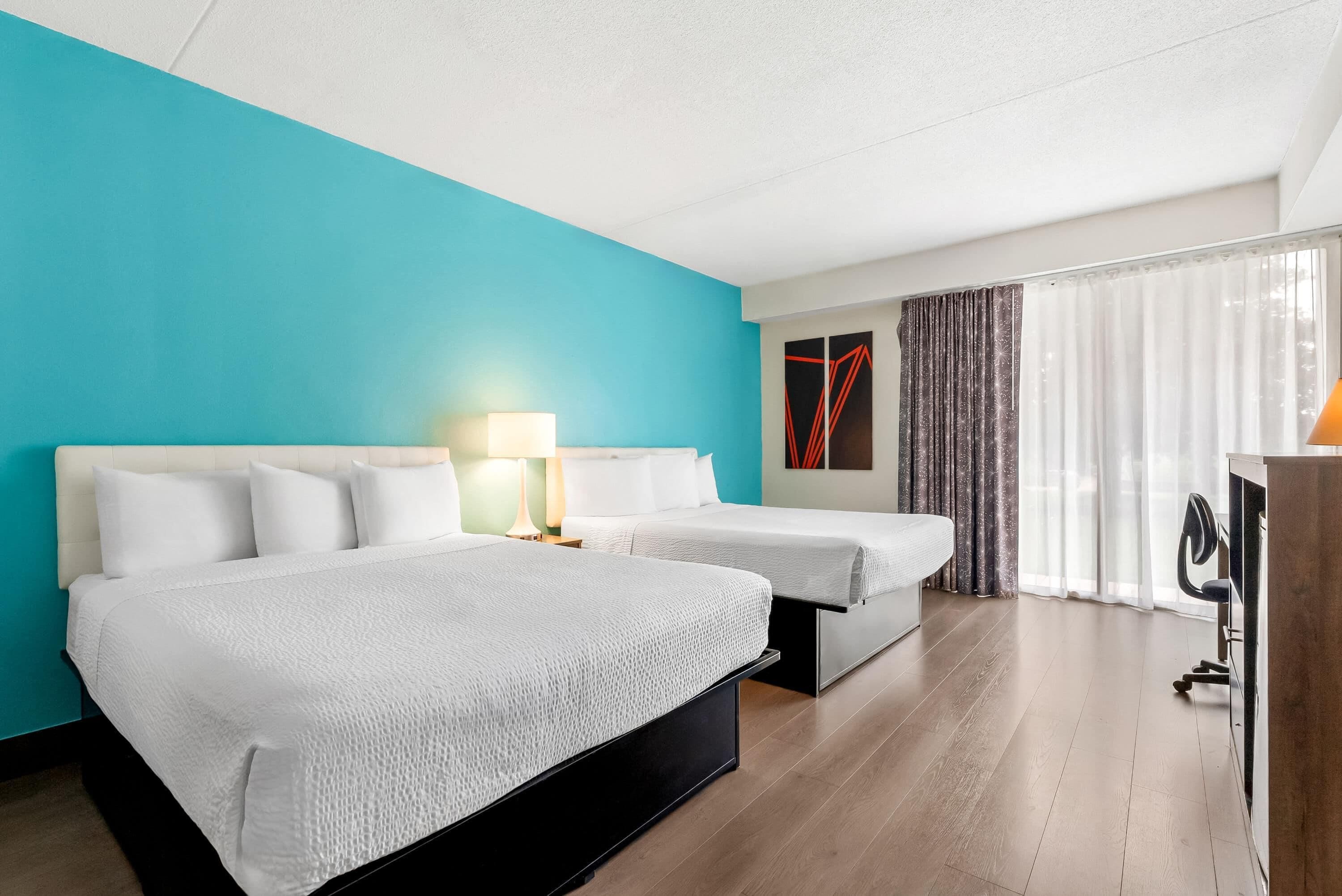 A hotel room with two beds, a desk, a lamp, and a view of the outdoors.