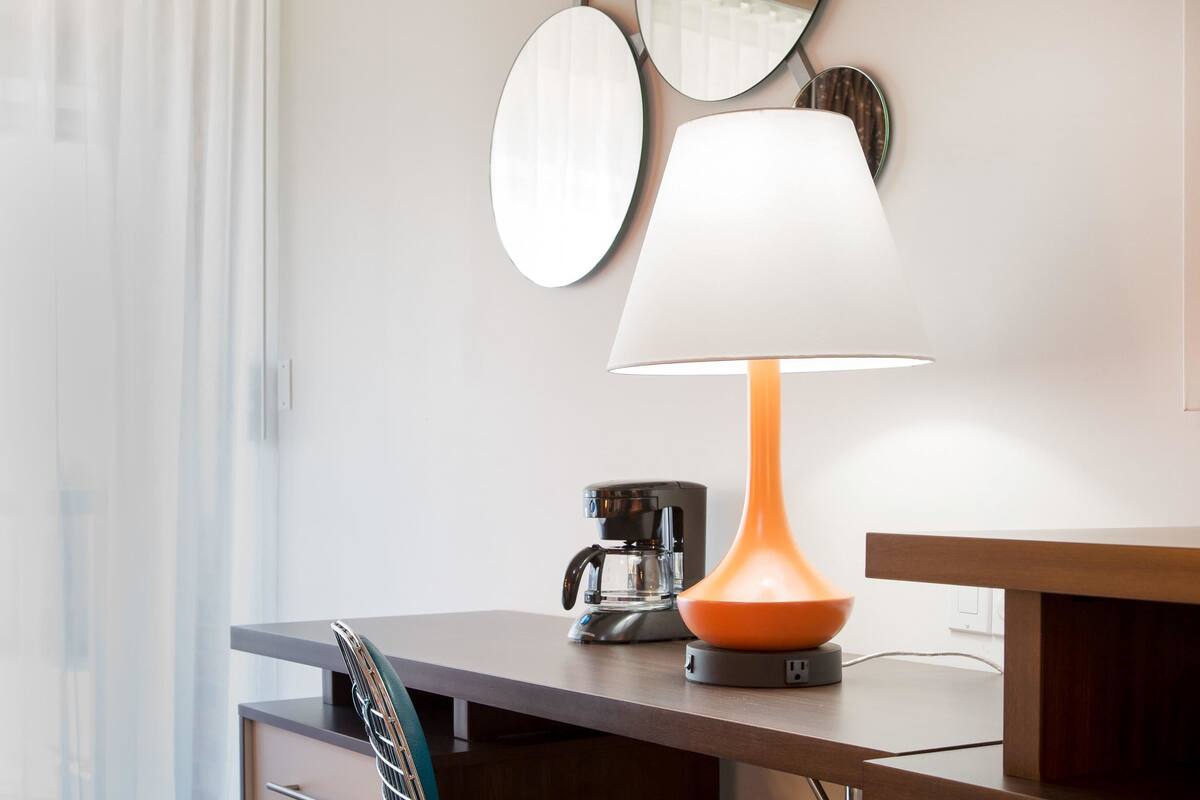 Une chambre moderne avec un bureau en bois, une machine à café et une lampe dont l’abat-jour est orange.