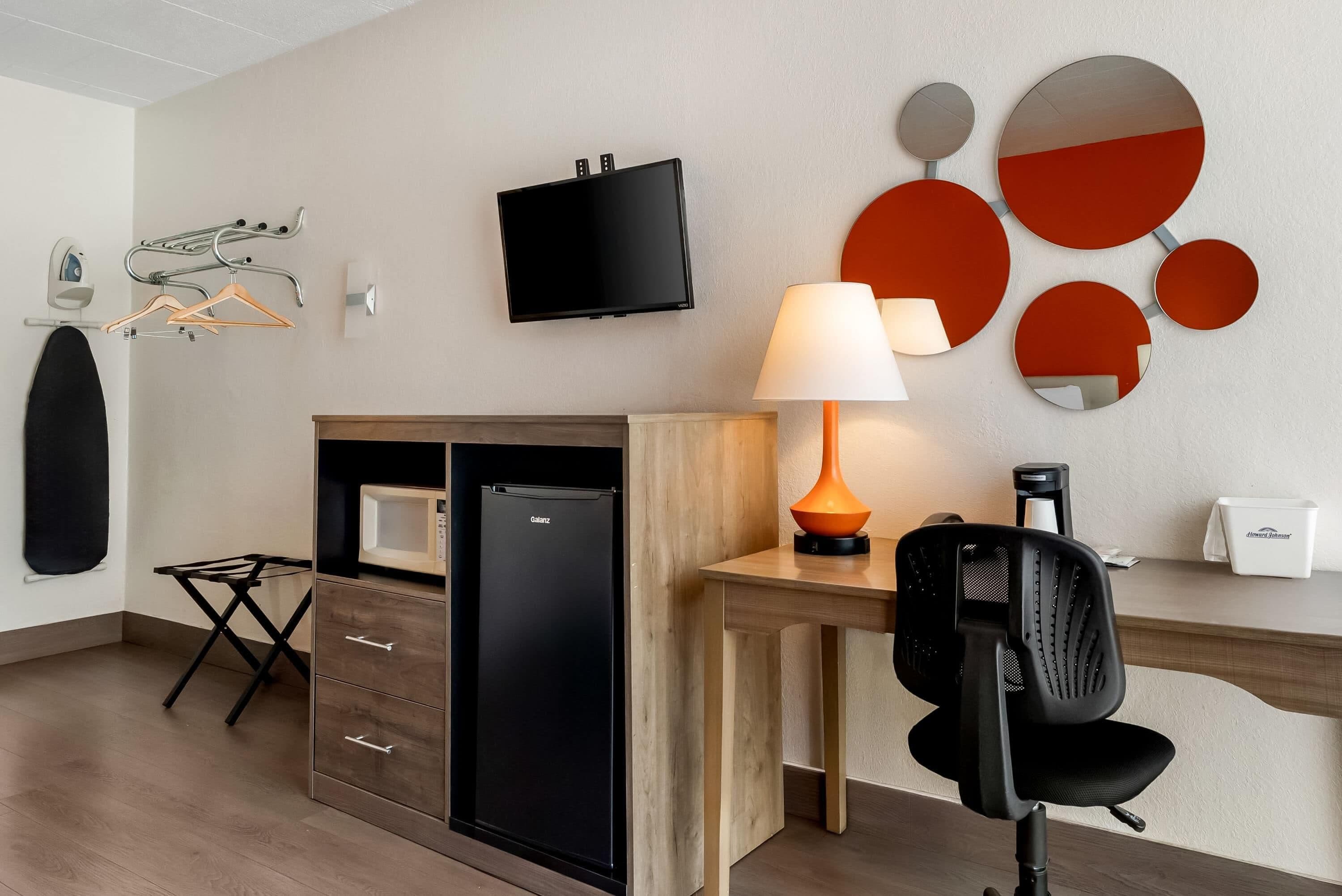 A hotel room with a desk, a chair, a television, a lamp, a mirror, and a minibar.