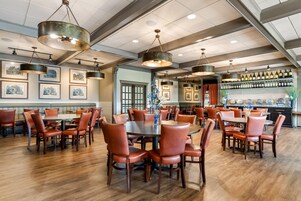 A restaurant interior with multiple dining tables and chairs, a bar area with wine bottles, and framed pictures on the walls.