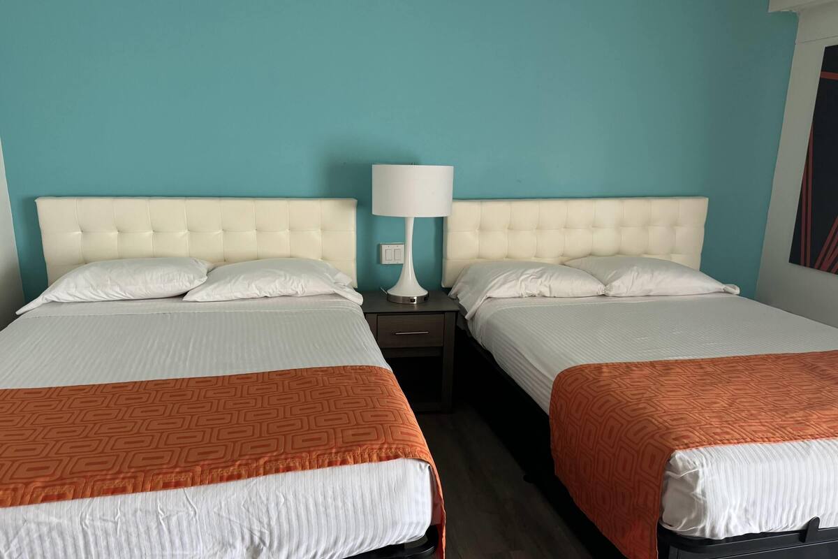 Une chambre d’hôtel avec deux lits, chacun étant équipé de draps blancs et d’une couverture à motifs orange. Une lampe blanche est posée sur une table de chevet entre les deux lits.