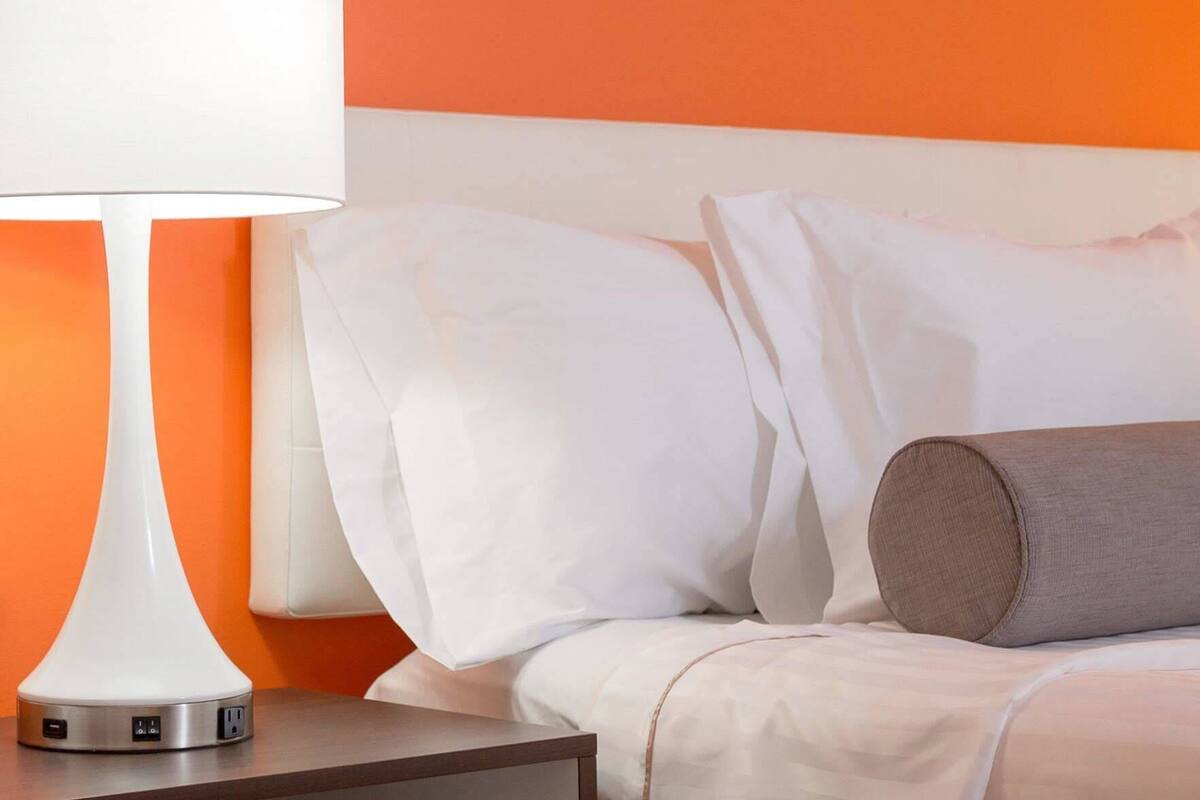 Un lit bien fait, avec des draps blancs, un oreiller marron et une lampe blanche posée sur une table de chevet.