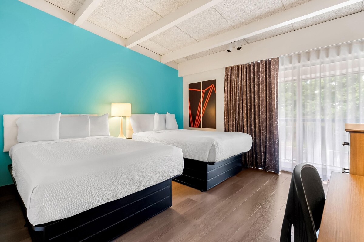 A hotel room with two beds, a desk, a chair, a lamp, and a large window with curtains.