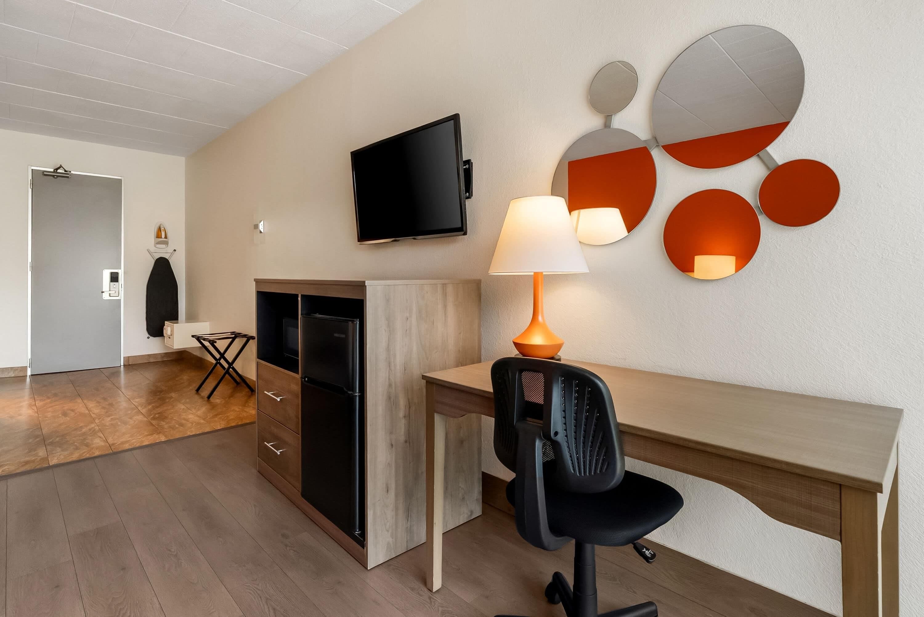 Et moderne hotelværelse med et fladskærms-TV, et skrivebord med stol, en lampe og et spejl monteret på væggen.