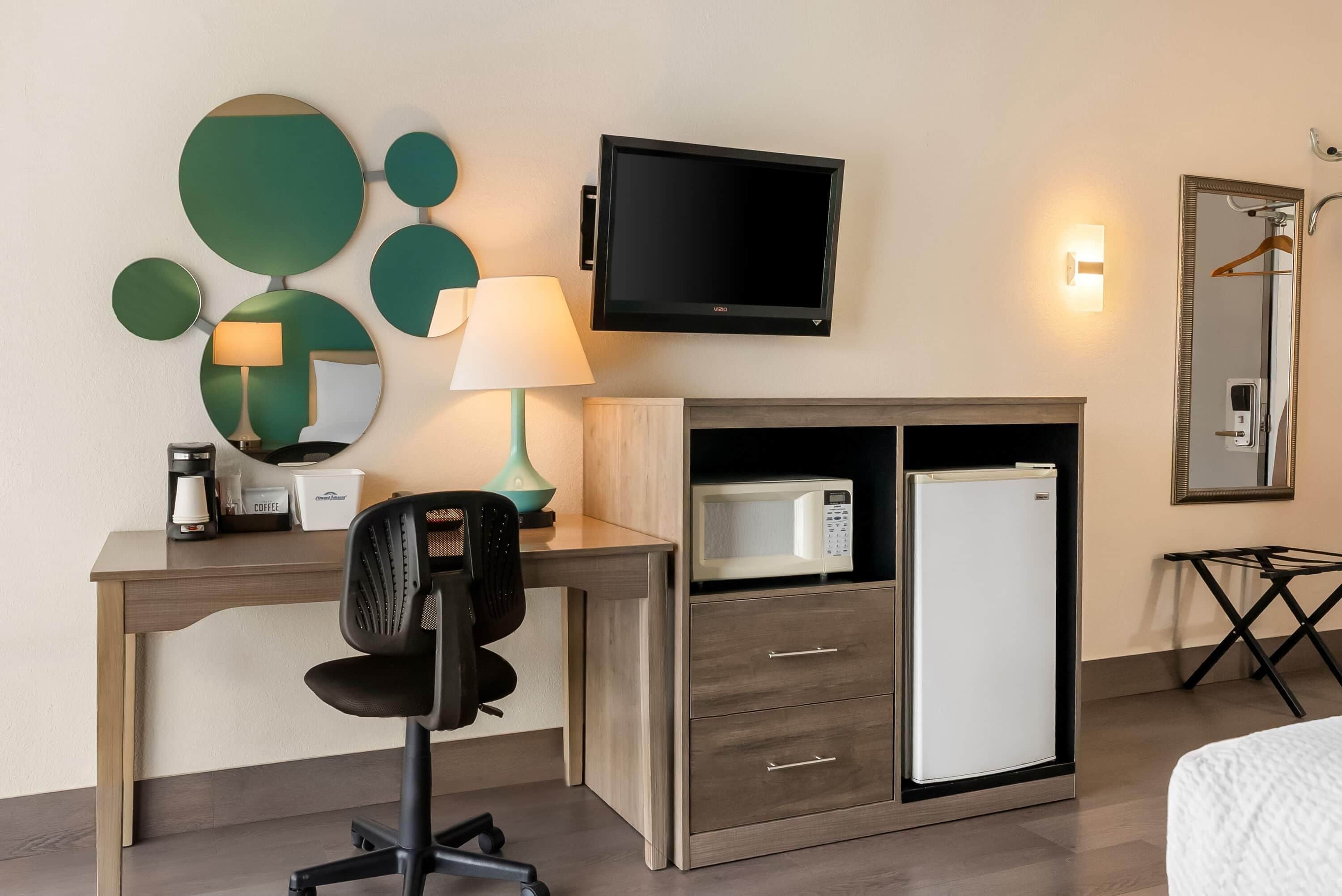 A hotel room with a desk, a chair, a television, a microwave, a refrigerator, a mirror, and a bedside table.