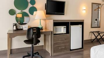 A hotel room with a desk, a chair, a television, a microwave, a refrigerator, a mirror, and a bedside table.