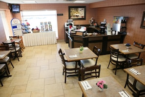 A restaurant interior with multiple tables and chairs, a counter with a coffee machine, and a television mounted on the wall.