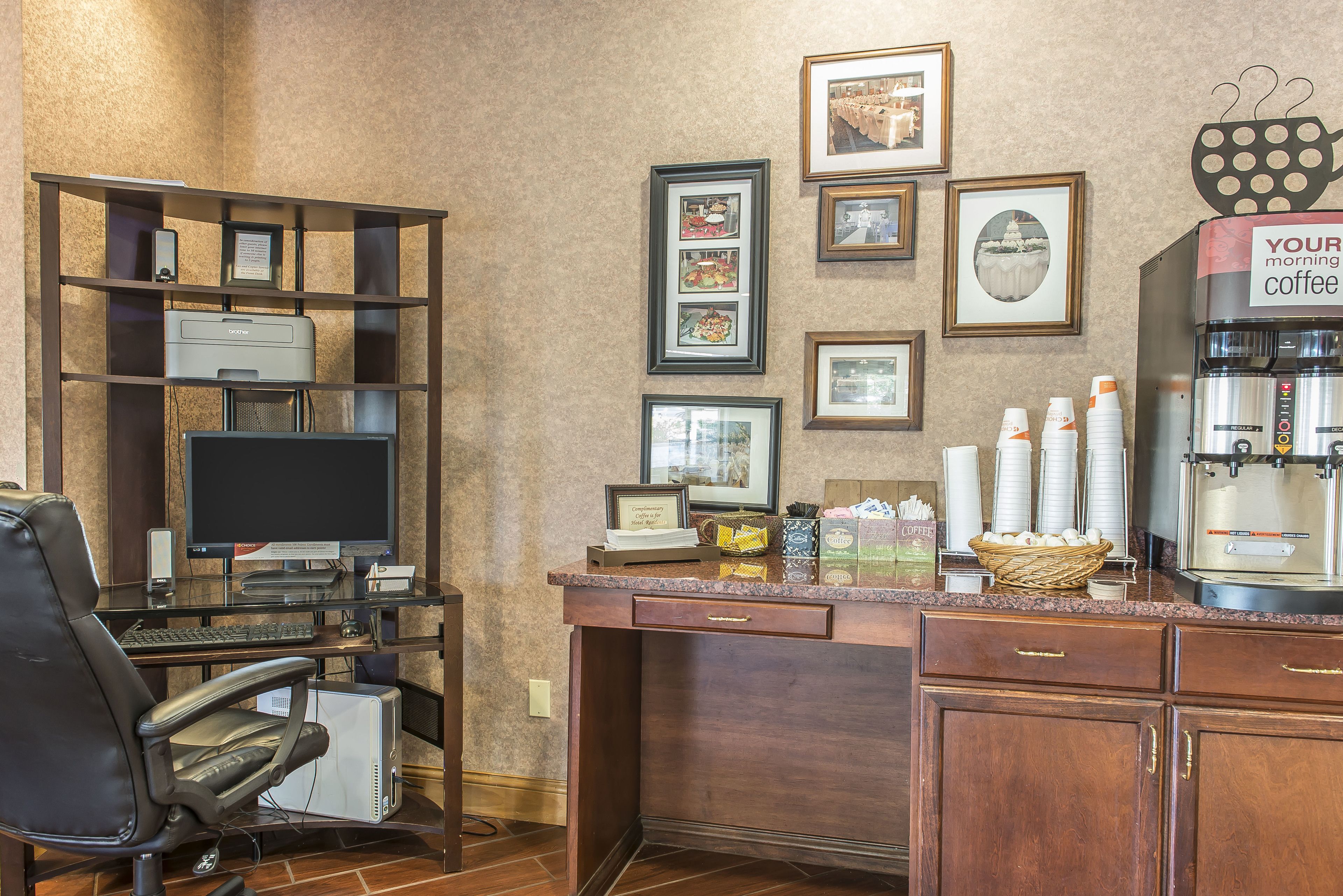 A hotel office with a computer desk, chair, printer, framed pictures on the wall, and a coffee station.