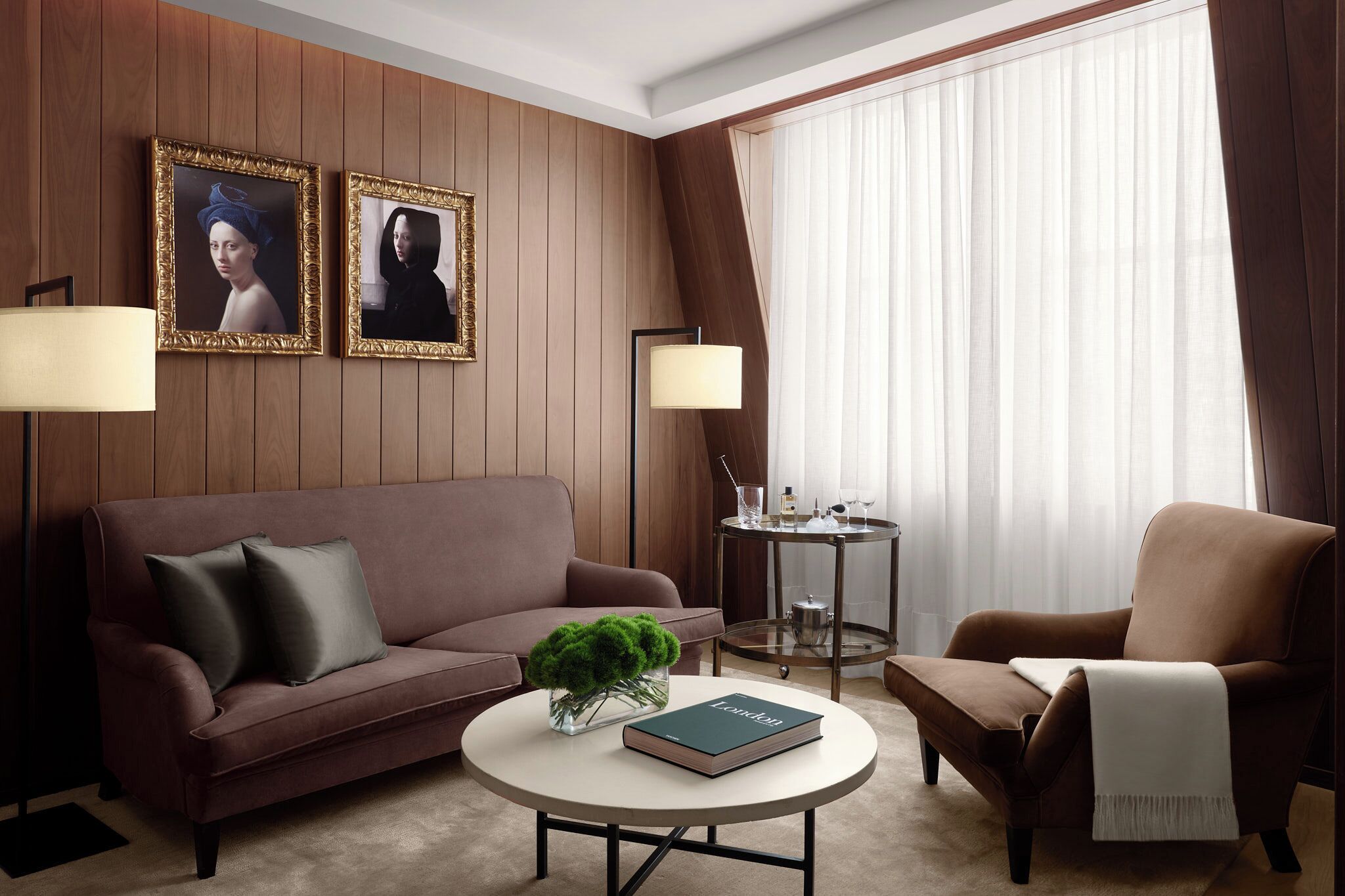 Une chambre d’hôtel moderne avec un canapé en cuir marron, une table basse ronde sur laquelle reposent un livre et une plante, une table d’appoint avec une lampe, et deux tableaux encadrés accrochés au mur.