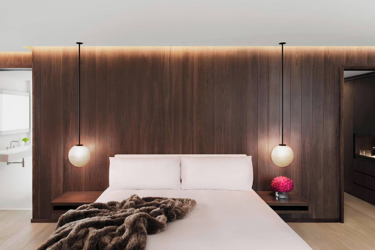 Un dormitorio moderno con una cama grande, dos lámparas colgantes, un panel de madera en la pared y una mesita de noche con un jarrón de flores.