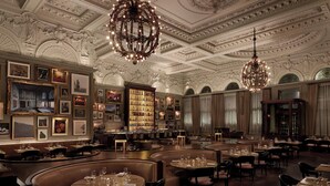 A luxurious dining area with ornate ceiling details, chandeliers, and a bar area with framed pictures. - The London EDITION (London)