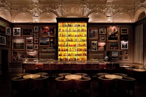 A bar area with a large illuminated liquor display, multiple framed pictures on the walls, and several round tables with chairs. - The London EDITION (London)
