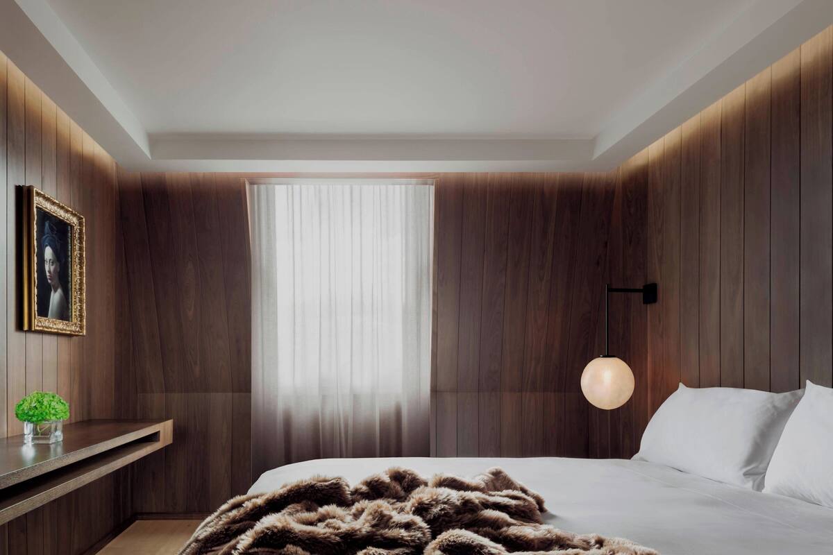 Un dormitorio moderno con una cama grande, paredes de paneles de madera, una mesita de noche con una lámpara y una ventana con cortinas traslúcidas.