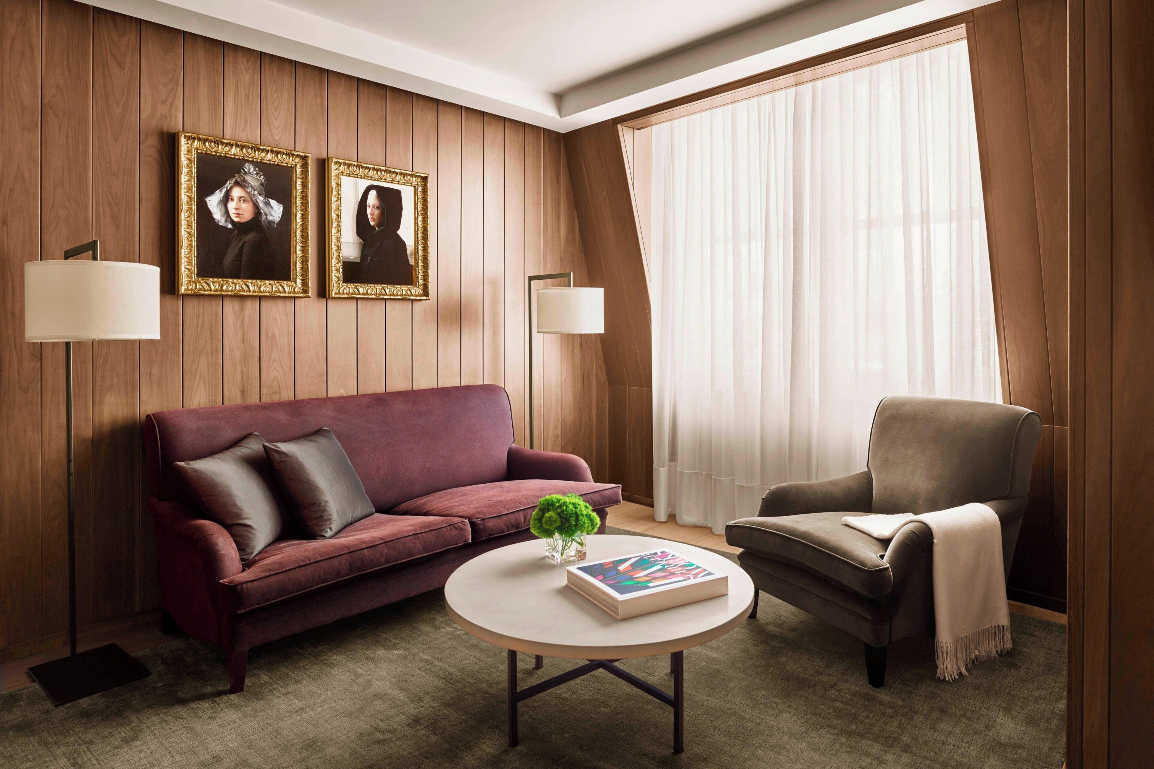 A room with a wooden paneled wall, a red sofa, a grey armchair, a round coffee table with a book and a plant, and two wall-mounted lamps.