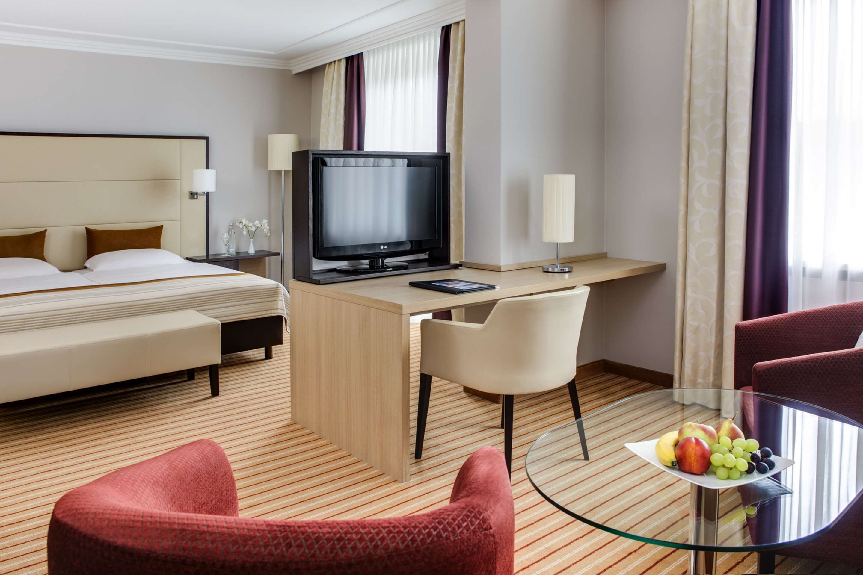 Une chambre d’hôtel avec un lit, un bureau avec une télévision, une table en verre avec des fruits et deux chaises.