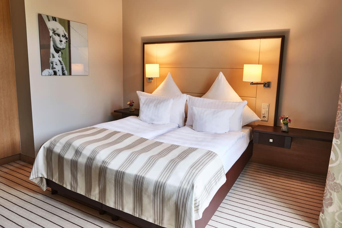 A hotel room with a large bed, two bedside tables, a headboard with integrated lighting, a framed artwork on the wall, and a wooden floor.