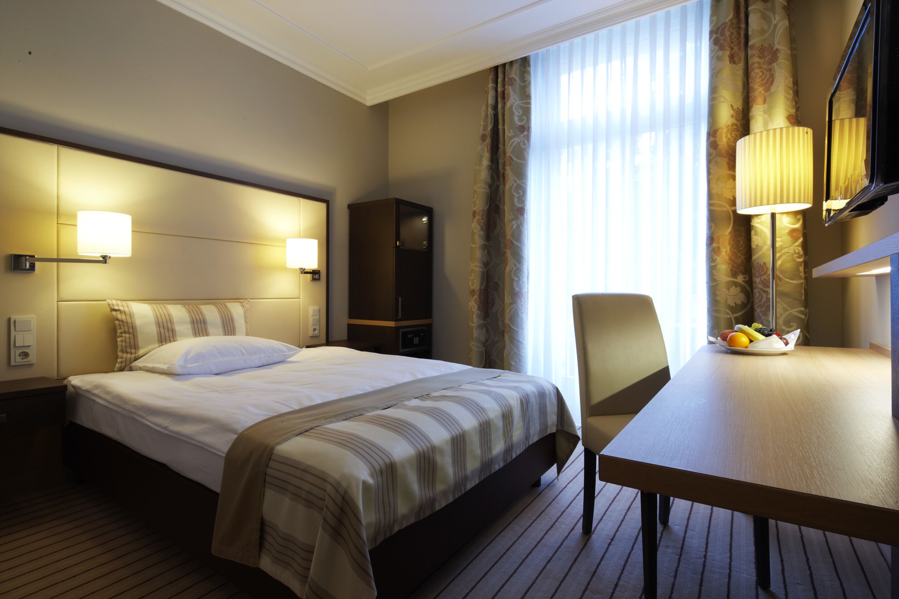 A hotel room with a bed, bedside lamps, a desk with a fruit bowl, and a mirror.
